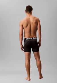 Calzoncillos bóxer negros con una cintura texturizada que presenta "CALVIN KLEIN" en blanco, vistos desde atrás, sobre un fondo gris neutro.