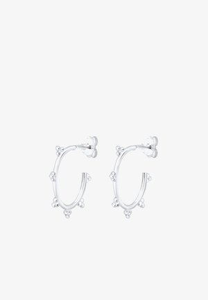 Boucle d'oreille créole en argent avec de petits détails en perles, portée à l'oreille gauche d'une personne aux cheveux châtain rouge tirés en arrière.