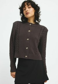 Brun kabelstickad cardigan med ribbad fåll, prydd med silverknappar och texturerade ärmar. Kort modell med rund halsringning.