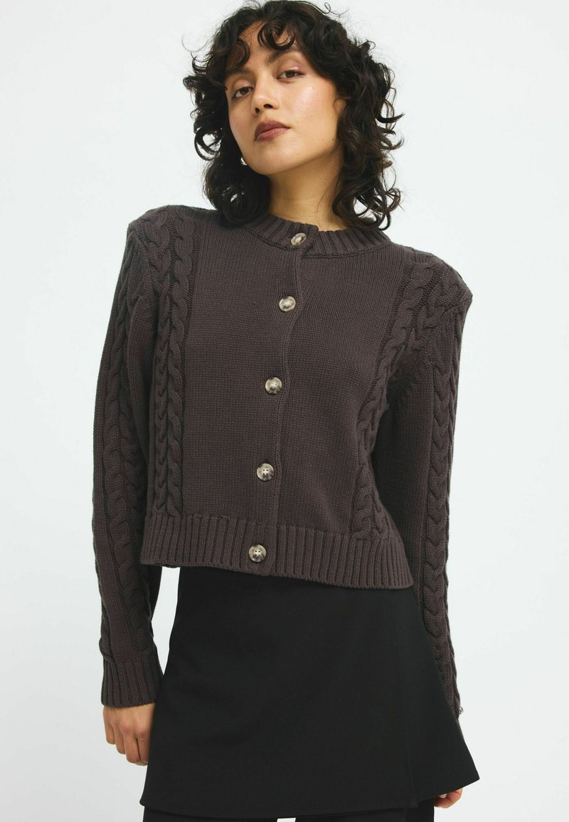 Brun kabelstickad cardigan med ribbad fåll, prydd med silverknappar och texturerade ärmar. Kort modell med rund halsringning.