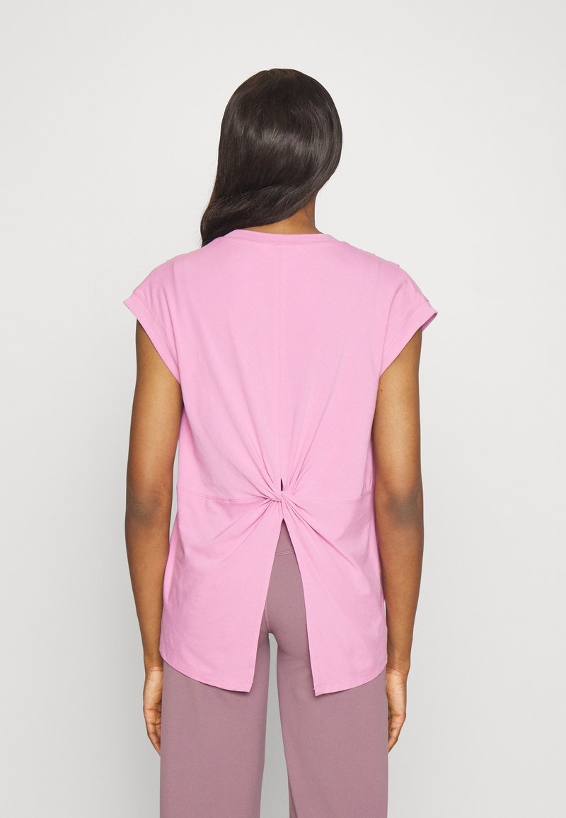 Femme portant un haut rose sans manches avec un nœud tordu et une fente dans le dos, associé à un pantalon large mauve, sur fond uni.