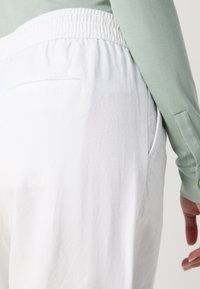 Personne portant un pantalon blanc à taille élastique et un haut vert clair à manches longues, visible de la taille vers le bas avec la main droite visible.