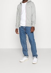 Felpa grigia con zip, cappuccio, tasche frontali e dettagli rossi, abbinata a jeans blu a gamba dritta e sneakers bianche.