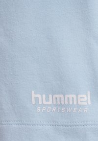 Hellblaues Baumwoll-Sportbekleidungsstück mit geripptem Textur und weißem, gedrucktem Logo mit der Aufschrift "hummel SPORTSWEAR" am unteren Ende. Einfaches Design.