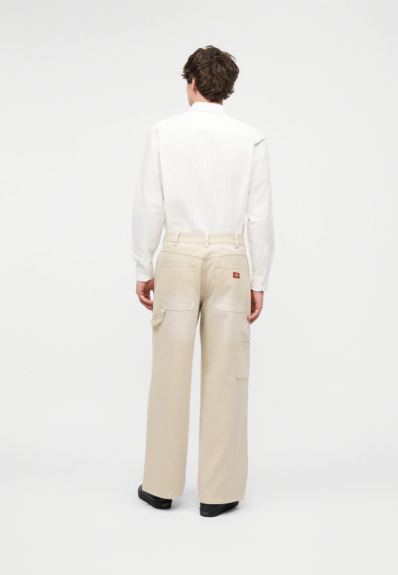 Homme portant un pantalon beige à jambes larges avec poches plaquées et une chemise blanche à manches longues, debout, de dos sur fond blanc.