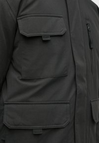 Dunkelgraue Jacke mit mehreren Brusttaschen, einer Reißverschlusstasche und einer glatten Textur. Das Design umfasst verstärkte Nähte und eine moderne Passform.