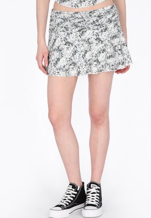 Kvinde iført en kort sort-hvid blomsterprint nederdel og sorte høje sneakers på en hvid baggrund.