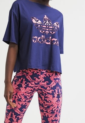 Kobieta nosząca granatowy t-shirt Adidas z różowym kwiatowym logo oraz dopasowane legginsy w granatowym i różowym kwiatowym wzorze.