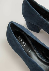 Zapatos de tacón alto de ante azul marino con puntera cerrada, que presentan una superficie texturizada y un tacón cuadrado. En el interior tienen un forro negro con la marca.