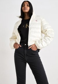 Casaco branco volumoso com colarinho alto e fecho de zip, usado sobre uma camisa preta com letras douradas. Combinado com jeans pretos. Textura suave.