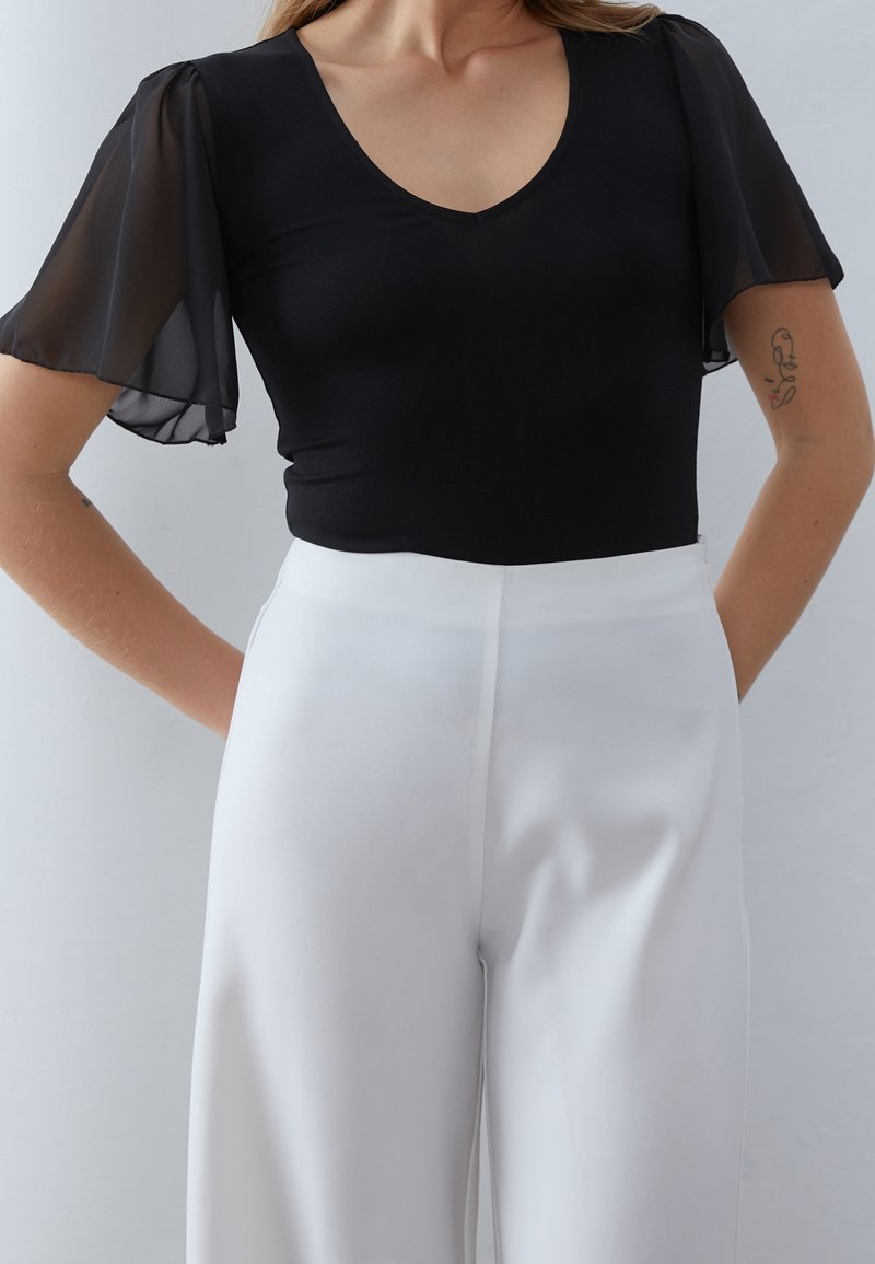 Femme portant un top noir à col en V avec des manches transparents flottantes et un pantalon blanc taille haute à jambes larges, debout les mains dans le dos.