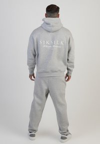 Hellgraues Hoodie- und Hosen-Set aus weichem Stoff. Das Hoodie hat ein großes weißes Logo auf dem Rücken. Lässige, lockere Passform mit gerippten Bündchen.