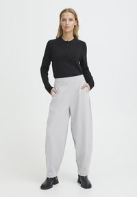 Polo noir à manches longues avec boutons, associé à un pantalon gris clair à jambes larges avec poches et texture lisse, porté avec des bottines noires.