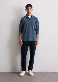 Blå langermet polo skjorte, myk stoff, avslappet passform; kombinert med mørke jeans og hvite sneakers, stående mot en nøytral bakgrunn.