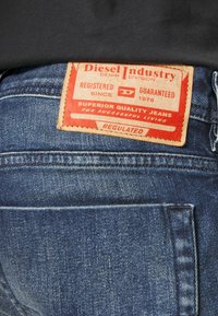 Farkut denim sinisellä tekstuurilla, joissa on keskeinen punainen nahkaleima, jossa lukee "Diesel Industry", kiinnitetty takavöihin.