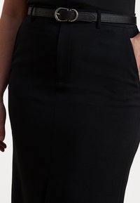 Μαύρη μολυβένια φούστα με λεία υφή, με εφαρμοστή σιλουέτα, και στενή μαύρη ζώνη με στρογγυλή ασημένια αγκράφα.