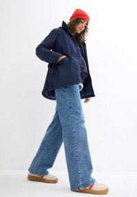 Giacca puffer blu navy con colletto alto, tasche frontali, indossata con jeans chiari a gamba larga e sneakers crema con accenti rossi, completata da un berretto rosso.