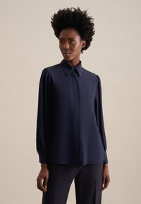 Blusa blu navy con colletto classico, maniche lunghe a sbuffo e texture liscia, dotata di chiusura con bottoni sulla parte anteriore. Design elegante e minimalista.