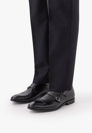 Czarne skórzane buty z podwójnym paskiem typu monk, noszone z czarnymi spodniami wizytowymi, pokazane od kostki w dół na białym tle.
