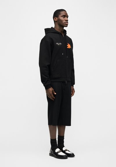 Νέος άνδρας που φοράει μαύρο φούτερ με φερμουάρ και πορτοκαλί λογότυπο πουλιού, μαύρα σορτς μέχρι το γόνατο, μαύρες κάλτσες και ασπρόμαυρα μοκασίνια, στέκεται πλαγίως.