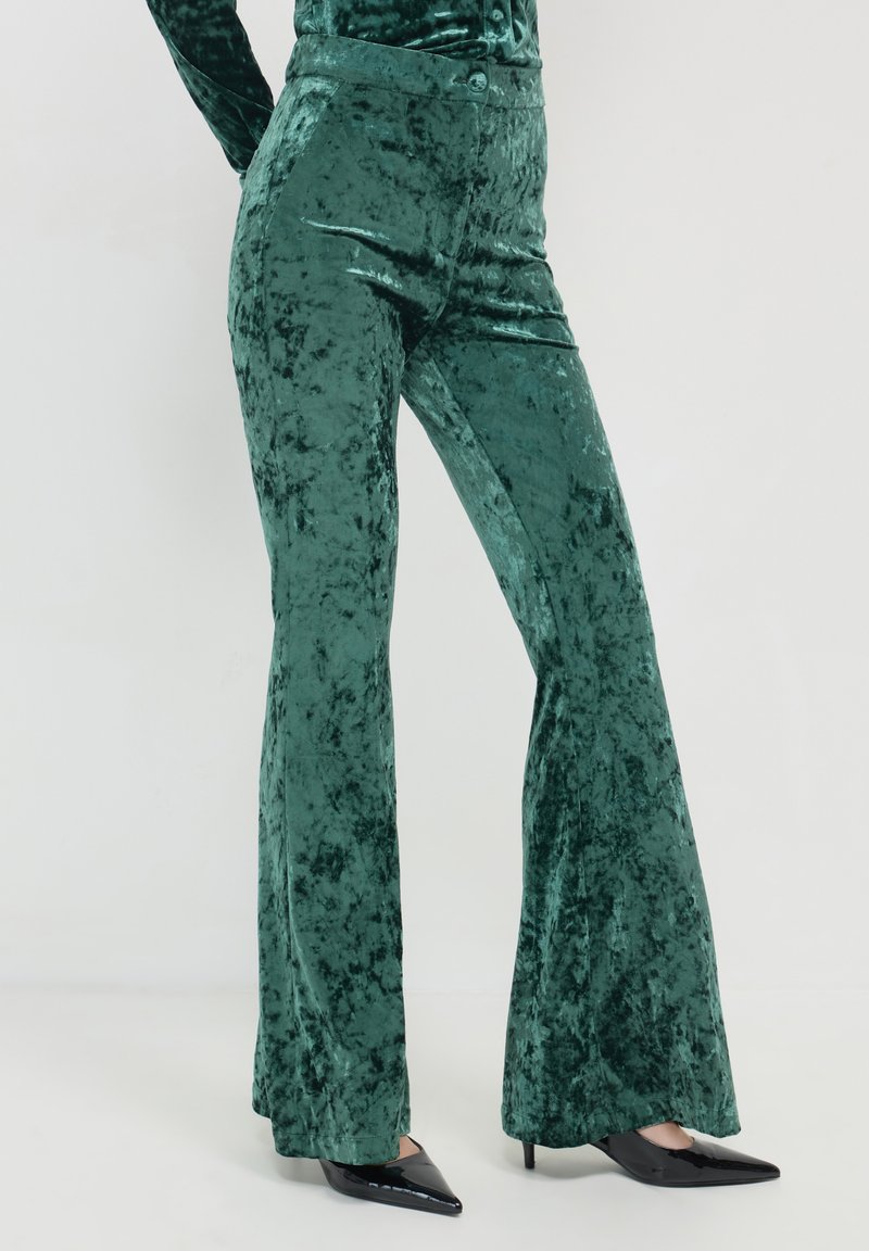 Pantaloni svasati in velluto verde con una texture morbida, caratterizzati da un design a vita alta e una leggera lucentezza, abbinati a tacchi neri appuntiti.