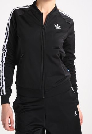 Osoba ubrana w czarny dres Adidas z białymi paskami na rękawach i spodniach, kurtka z zamkiem z przodu oraz logo na klatce piersiowej i udzie.