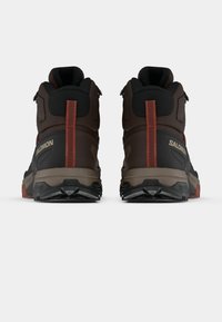 Καφέ ψηλά ορειβατικά παπούτσια με μαύρες λεπτομέρειες, υλικό με υφή, συγκολλημένες επικαλύψεις και κόκκινη ετικέτα στη φτέρνα. Διαθέτουν εξωτερική σόλα από καουτσούκ για πρόσφυση.