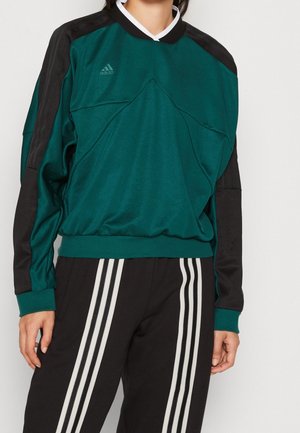 Person iført en teal og sort Adidas sweatshirt med en hvid V-hals krave og sorte bukser med tre hvide striber ned ad benene.