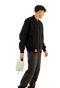 Chaqueta bomber negra con puños acanalados y una etiqueta naranja, combinada con pantalones gris oscuro. Sosteniendo una bolsa gris claro con un asa negra.