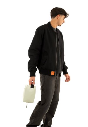 Chaqueta bomber negra con puños acanalados y una etiqueta naranja, combinada con pantalones gris oscuro. Sosteniendo una bolsa gris claro con un asa negra.