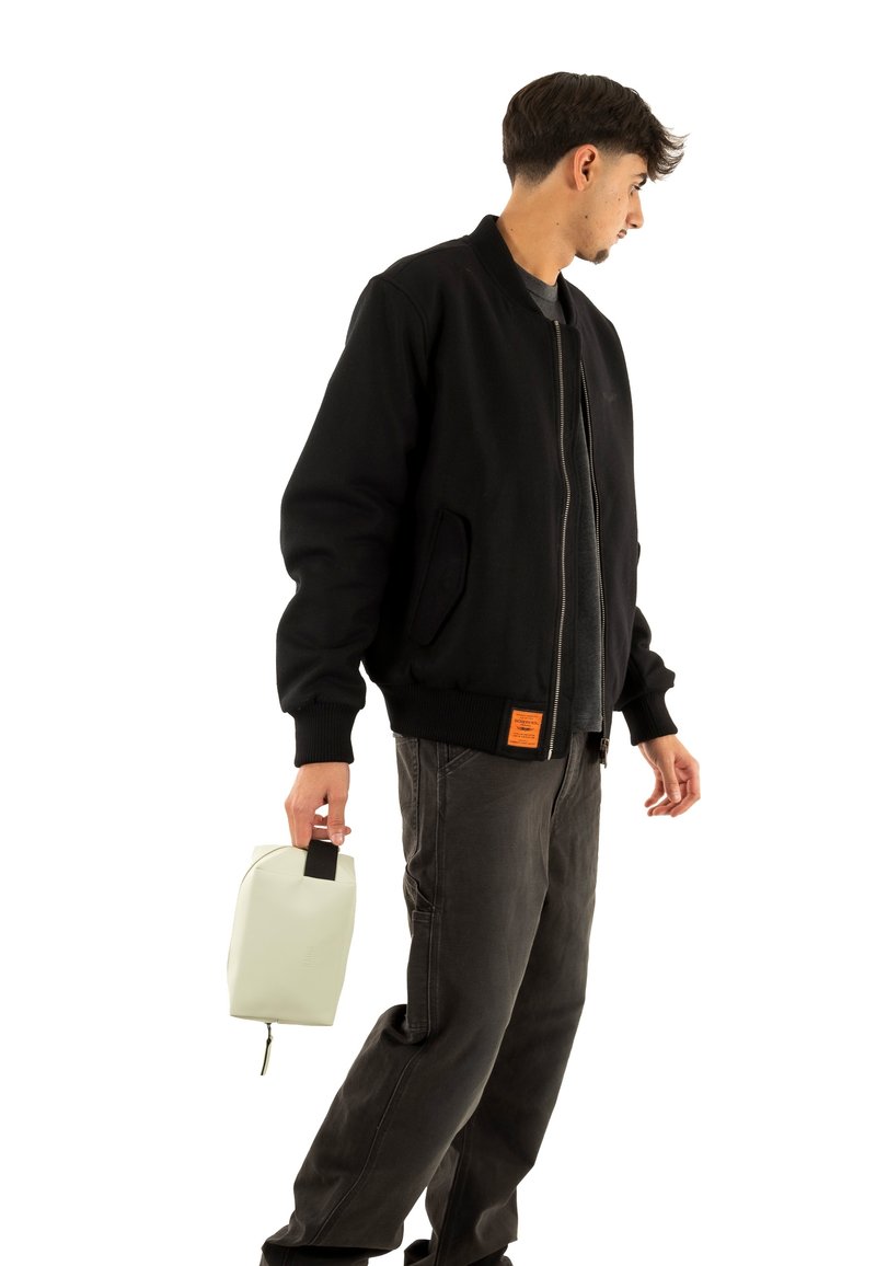 Chaqueta bomber negra con puños acanalados y una etiqueta naranja, combinada con pantalones gris oscuro. Sosteniendo una bolsa gris claro con un asa negra.
