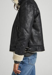 Veste en cuir noir avec un col et des poignets en fourrure, poche latérale zippée, et un design court avec un détail ceinturé à la taille.