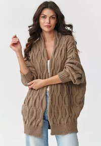 Brązowy sweter z dzianiny z teksturowanym wzorem w warkocze, otwarty z przodu, z luźnymi rękawami i ściągaczem na dole. Nosi się go na prostą bluzkę i dżinsy.
