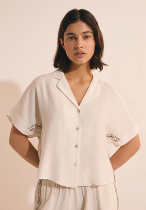 Młoda kobieta w luźnej, kremowej koszuli z krótkim rękawem z subtelnymi haftowanymi detalami, stoi na tle jednolitego beżowego tła.