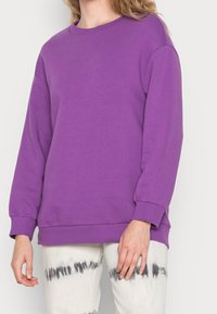 Sudadera morada con cuello redondo, mangas largas y puños acanalados, hecha de material suave; combinada con pantalones tie-dye.