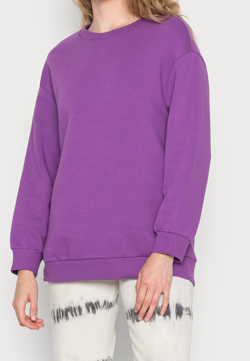 Sudadera morada con cuello redondo, mangas largas y puños acanalados, hecha de material suave; combinada con pantalones tie-dye.