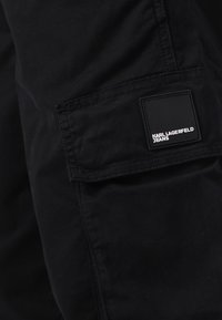 Fekete cargo nadrág sima textúrával, oldalsó zsebbel és téglalap alakú márkalábbal, "KARL LAGERFELD JEANS" felirattal. Egyszerű varrás részletek.