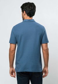 Mies selinpäin, yllään sininen lyhythihainen poolopaita ja tummat farkut, yksinkertaista vaaleaa taustaa vasten.