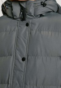 Giacca imbottita grigia con cappuccio. Presenta chiusura con cerniera, bottoni a pressione neri e cuciture orizzontali. Tessuto liscio, design leggero.