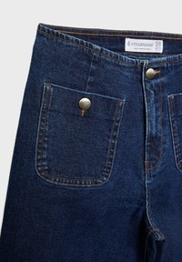 Pantalon en denim bleu foncé avec poches devant à rabat maintenues par des boutons métalliques et coutures orange visibles, étiquette de taille 38 à l'intérieur de la ceinture.