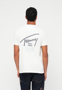 Λευκό βαμβακερό T-shirt με μπλε γραφικά κείμενα στην πλάτη, που περιλαμβάνουν το "Tommy" και ονόματα πόλεων. Στρογγυλή λαιμόκοψη, κοντά μανίκια, άνετη εφαρμογή.