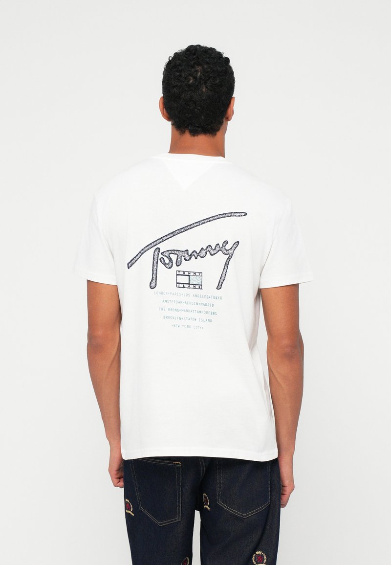 Λευκό βαμβακερό T-shirt με μπλε γραφικά κείμενα στην πλάτη, που περιλαμβάνουν το "Tommy" και ονόματα πόλεων. Στρογγυλή λαιμόκοψη, κοντά μανίκια, άνετη εφαρμογή.