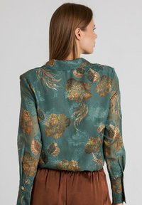 Blusa floral verde con tejido translúcido y bordado texturizado en oro y azul verdoso, mangas largas y cuello clásico.