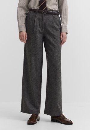 Person trägt graue, weit geschnittene Hose mit hoher Taille mit schwarzem Gürtel, braune Loafers, ein gestreiftes Hemd und eine gestreifte Krawatte.
