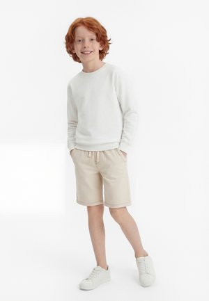 Rudovlasý chlapec stojí s rukami vreckách, má na sebe bielu mikinu, béžové šortky a biele tenisky na jednoliatom bielom pozadí.