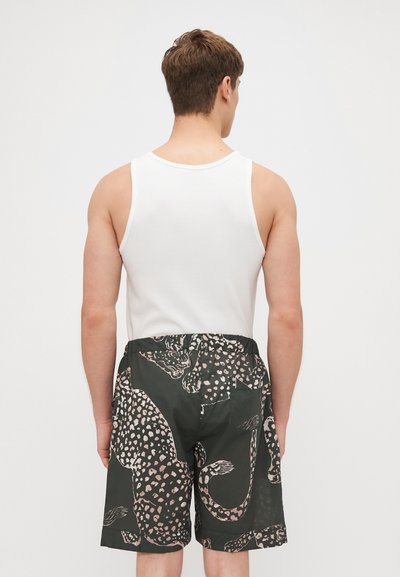 Maillots de bain pour hommes avec une base noire, présentant un imprimé léopard rose. Taille élastique et poche arrière unique. Porté avec un débardeur blanc.