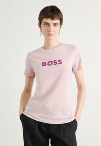 Šviesiai rožinė medvilninė marškinėliai su trumpomis rankovėmis, kuriuose krūtinės srityje ryškiai tamsiai violetinėmis raidėmis parašyta "BOSS".