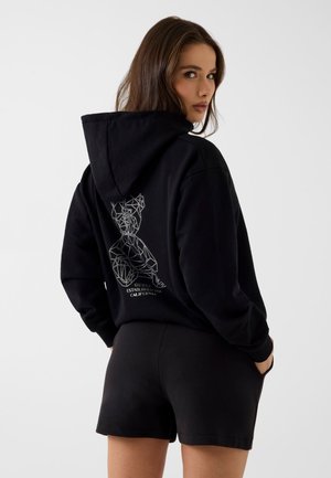 Frau mit langen braunen Haaren, die einen schwarzen Hoodie mit geometrischer Figur und Text auf dem Rücken trägt, kombiniert mit schwarzen Shorts, schaut über die Schulter.