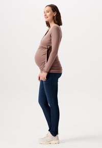 Graviditetstop i blødt, lysebrunt stof med en bindedetalje, parret med mørkeblå skinny jeans og lyse sneakers, set fra siden.