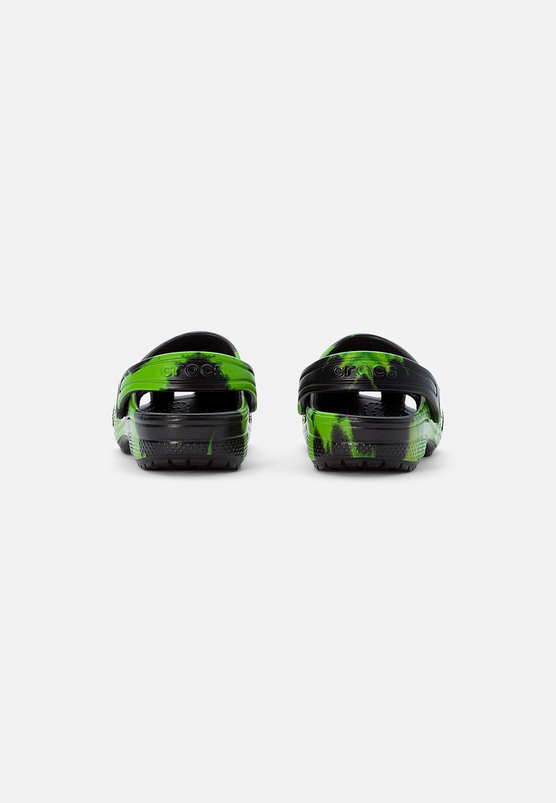 Un par de Crocs de tie-dye en negro y verde vistos desde atrás, mostrando las correas en el talón y las suelas texturizadas sobre un fondo blanco.